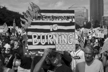 A protester at a No Kings march holds up a poster that is made to look like it is on fire that says "Democracy is burning."