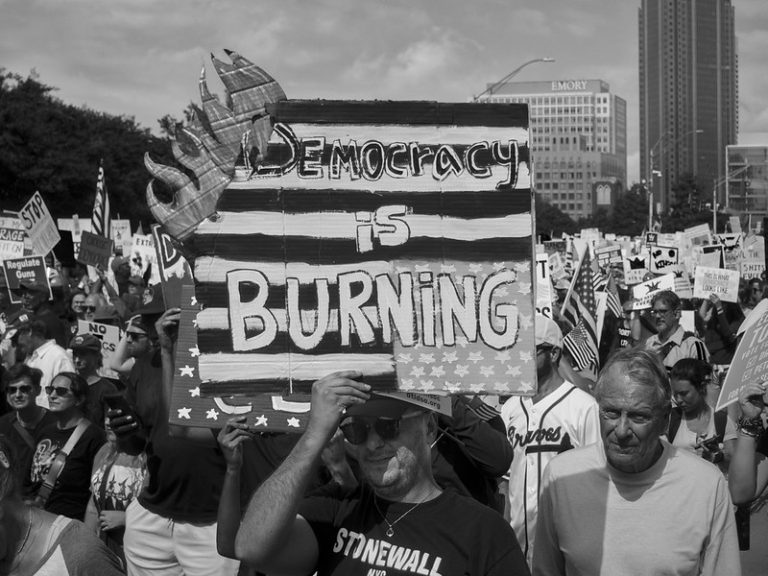 A protester at a No Kings march holds up a poster that is made to look like it is on fire that says "Democracy is burning."